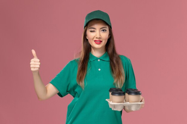 Front view female courier in green uniform and cape holding delivery coffee cups winking on pink wall service uniform delivery job