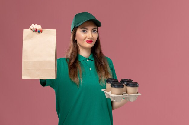 Front view female courier in green uniform and cape holding food package and delivery coffee cups on pink desk service uniform delivery job