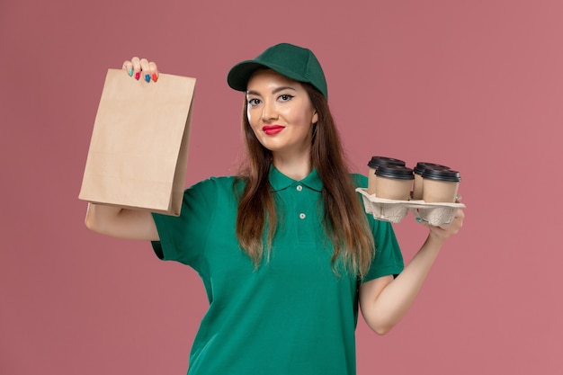 Front view female courier in green uniform and cape holding food package and delivery coffee cups smiling on pink desk service uniform delivery job