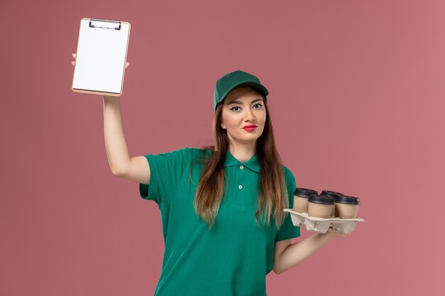 緑のユニフォームと薄ピンクの壁にメモ帳と配達コーヒーカップを保持しているケープの正面図の女性の宅配便