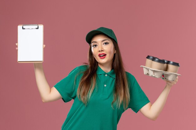 緑の制服とピンクの壁にメモ帳と配達のコーヒーカップを保持しているケープの正面図の女性の宅配便制服配達仕事の女の子