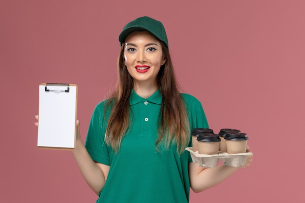 Front view female courier in green uniform and cape holding notepad and delivery coffee cups on pink wall service uniform delivery job worker