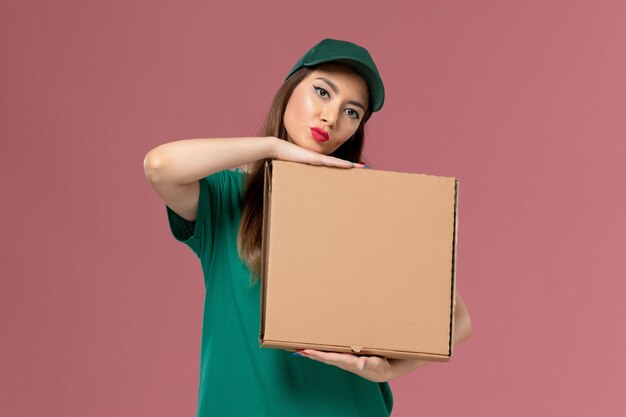 Женщина-курьер в зеленой униформе, держащая коробку с едой на светло-розовой стене, служба доставки униформы