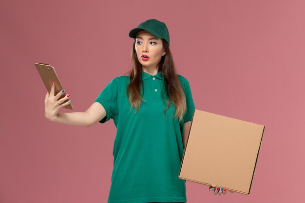 Front view female courier in green uniform holding food box and notepad on pink wall company service uniform delivery job work