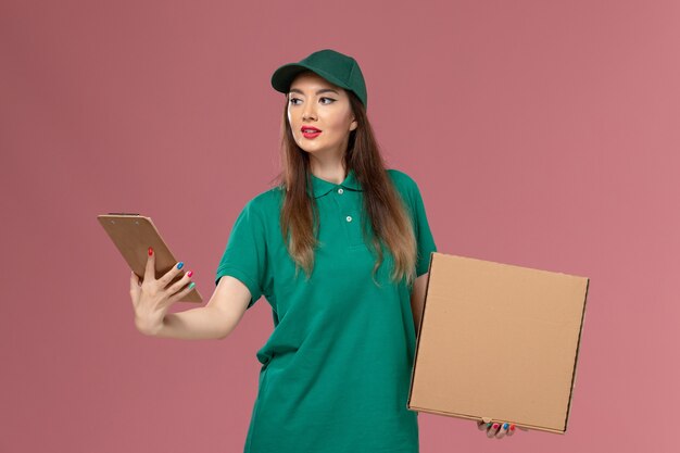 Front view female courier in green uniform holding food box and notepad on the pink wall company service worker uniform delivery job work