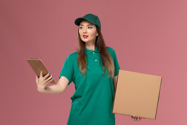 Вид спереди женщина-курьер в зеленой форме, держащая коробку с едой и блокнот на розовой стене, сервисный работник компании, униформа, доставка, работа