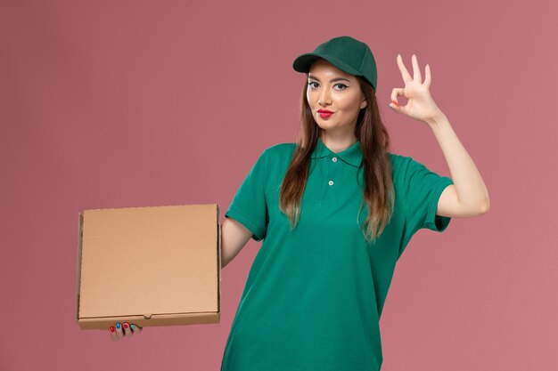 ピンクの壁にフードボックスを保持している緑の制服を着た正面図の女性宅配便仕事サービス制服配達作業