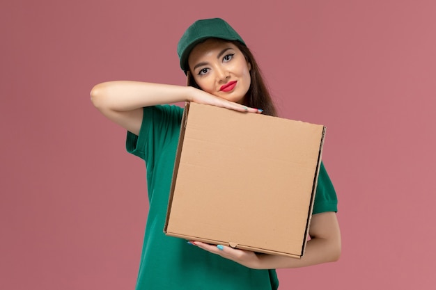 Corriere femminile di vista frontale in scatola di cibo della tenuta dell'uniforme verde con il sorriso sulla consegna uniforme di servizio dell'operaio di lavoro della parete rosa-chiaro