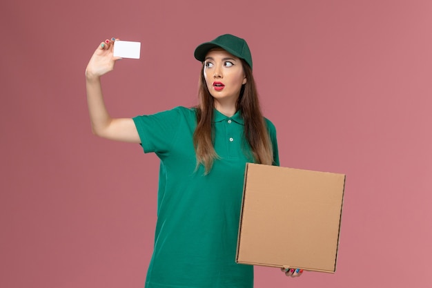 Вид спереди женщина-курьер в зеленой форме, держащая коробку для доставки еды и карточку на розовой стене, сервисная форма компании, работа по доставке