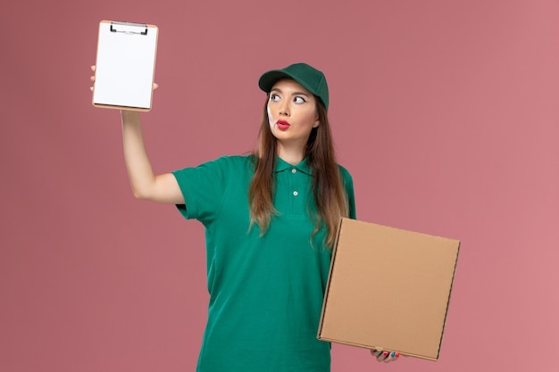 Front view female courier in green uniform holding food delivery box and notepad on the pink desk company service worker uniform delivery job