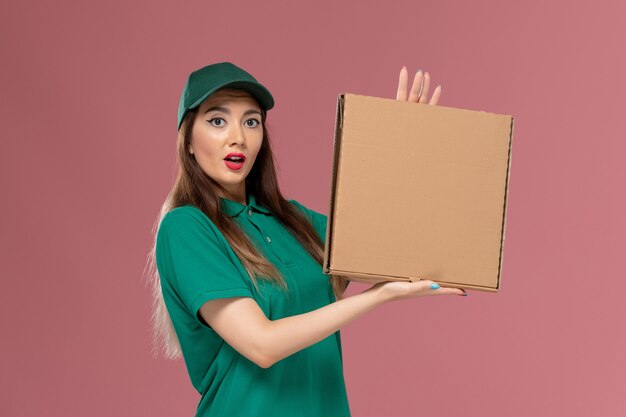 Front view female courier in green uniform holding food delivery box on the pink wall job worker service uniform delivery girl company