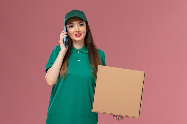 Front view female courier in green uniform holding food delivery box and talking on the phone on pink wall company service uniform delivery