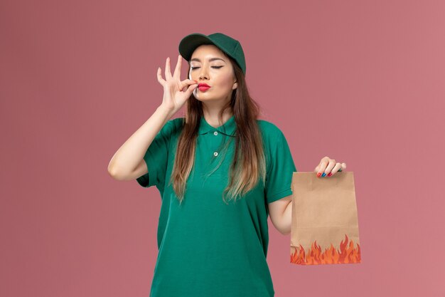 Front view female courier in green uniform holding food package on the light pink wall service uniform delivery