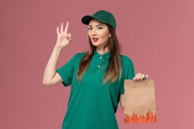 Вид спереди женщина-курьер в зеленой форме, держащая пакет с едой на розовой стене, доставка рабочей одежды