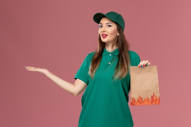 Front view female courier in green uniform holding food package on pink wall service uniform job delivery