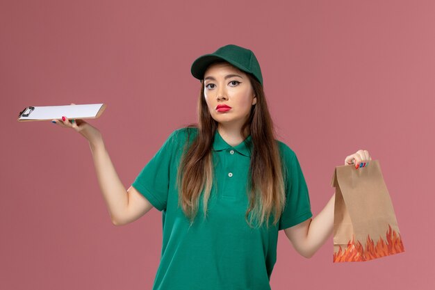 ピンクの壁のサービスの制服の配達作業の仕事でメモ帳と食品パッケージを保持している緑の制服の正面図の女性の宅配便