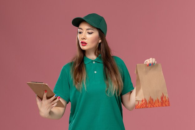 ピンクの壁のサービス制服配達労働者の仕事でメモ帳と食品パッケージを保持している緑の制服の正面図女性宅配便