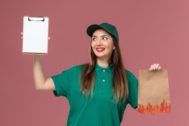 Front view female courier in green uniform holding notepad and food package on pink wall service worker uniform delivery job