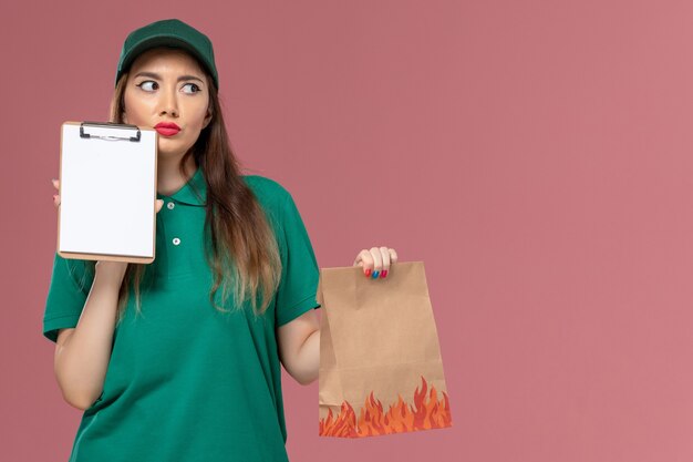 분홍색 벽 서비스 유니폼 배달 작업 노동자에 생각하는 메모장과 음식 패키지를 들고 녹색 제복을 입은 전면보기 여성 택배