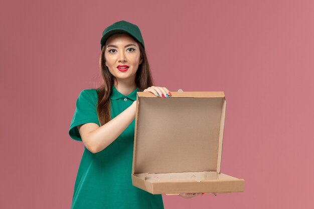 Front view female courier in green uniform holding and opening empty food box on the pink wall job service uniform delivery worker