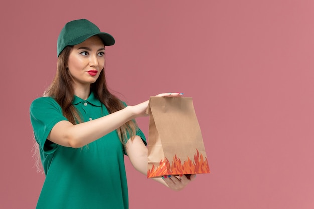 Женщина-курьер в зеленой форме, держащая бумажный пакет с едой на светло-розовой стене, служба доставки униформы