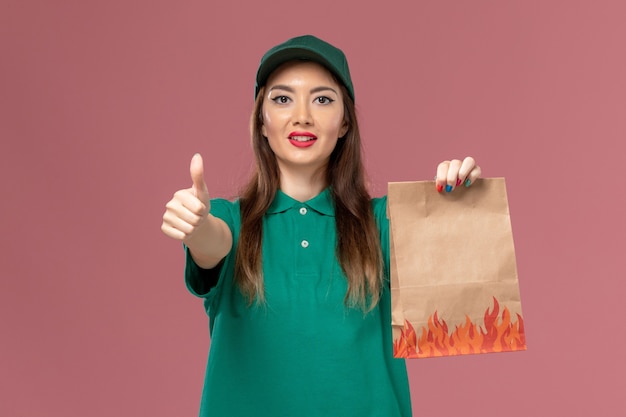 Вид спереди женщина-курьер в зеленой форме, держащая бумажный пакет с едой на розовой стене, служба доставки униформы