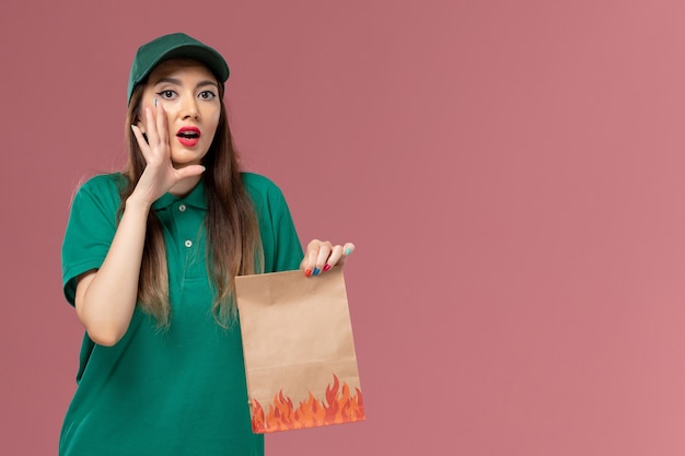 ピンクの壁のサービスの制服配達の仕事で紙の食品パッケージを保持している緑の制服の正面図の女性の宅配便