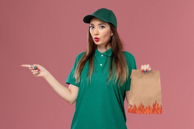 Front view female courier in green uniform holding paper food package on pink wall service uniform delivery