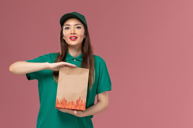 Вид спереди женщина-курьер в зеленой форме, держащая бумажный пакет с едой на розовой стене, сервисная форма, доставка работника