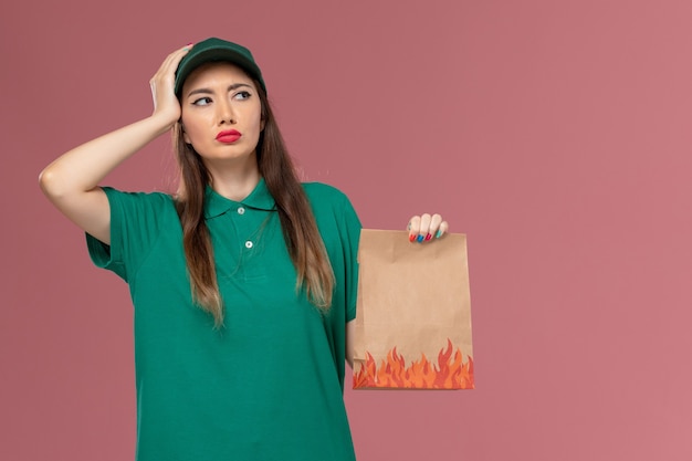 Front view female courier in green uniform holding paper food package thinking on pink wall service uniform delivery