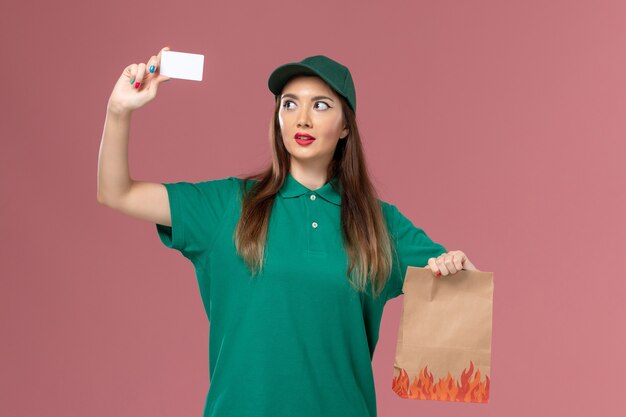 Front view female courier in green uniform holding white card and food package on pink wall service uniform delivery job worker