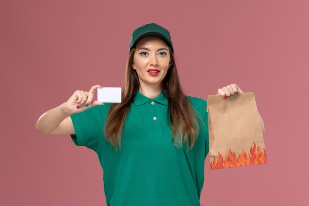 Вид спереди женщина-курьер в зеленой форме держит белую карточку и пакет с едой на розовой стене.