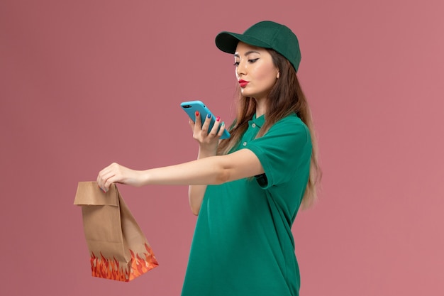 Front view female courier in green uniform taking photo of food package on pink wall service uniform delivery job worker