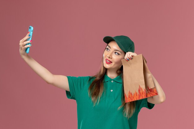 Front view female courier in green uniform taking selfie and holding food package on pink wall service uniform delivery job worker