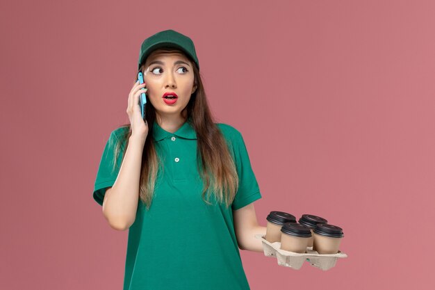 Front view female courier in green uniform talking on the phone and holding delivery coffee cups on light pink wall service uniform delivery job