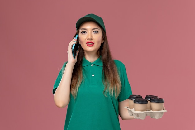 Front view female courier in green uniform talking on the phone and holding delivery coffee cups on pink desk service uniform delivery job