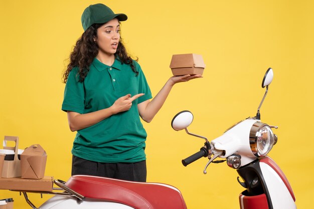 Front view female courier in green uniform with little food package on yellow background work color job delivery food service worker