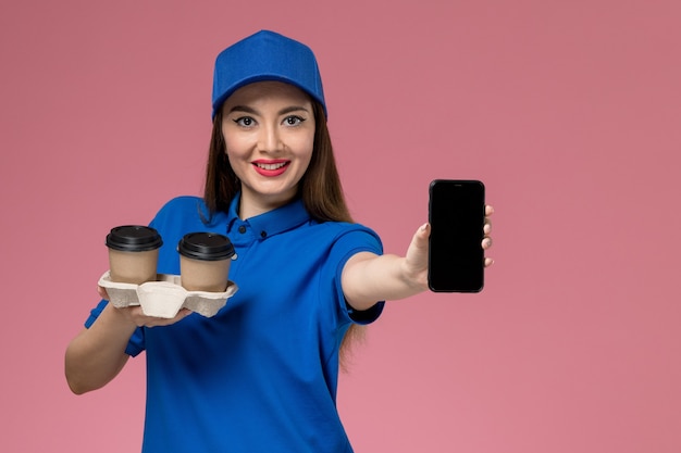 Бесплатное фото Женский курьер в синей униформе и плаще, держащий доставки кофе и телефон, улыбается на розовой стене
