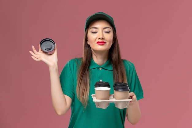 무료 사진 녹색 유니폼과 케이프 핑크 벽 서비스 유니폼 배달 작업에 냄새 배달 커피 컵을 들고 전면보기 여성 택배