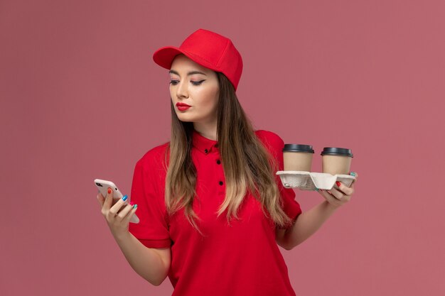 무료 사진 분홍색 배경 서비스 배달 유니폼에 그녀의 전화를 사용하여 배달 커피 컵을 들고 빨간 제복을 입은 전면보기 여성 택배