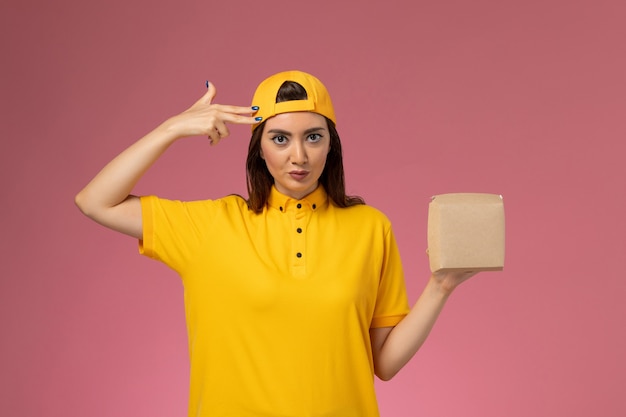 Вид спереди женщина-курьер в желтой форме и накидке с маленьким пакетом еды для доставки на светло-розовой стене.