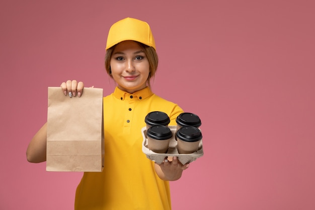 Бесплатное фото Вид спереди женщина-курьер в желтой униформе, желтой накидке, держащей кофейные чашки с едой на розовом фоне, цвет униформы доставки