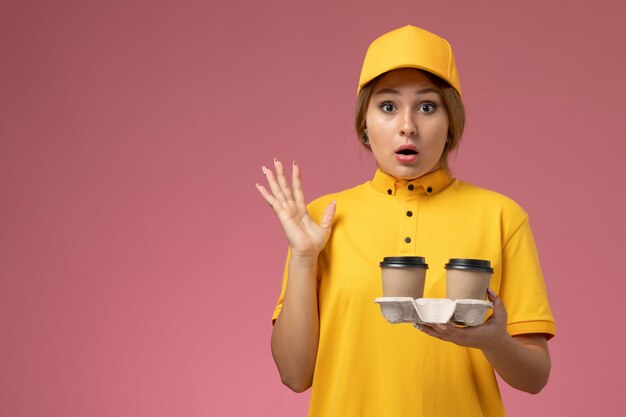 Бесплатное фото Вид спереди женщина-курьер в желтой униформе с желтым плащом держит пластиковые кофейные чашки на розовом фоне.