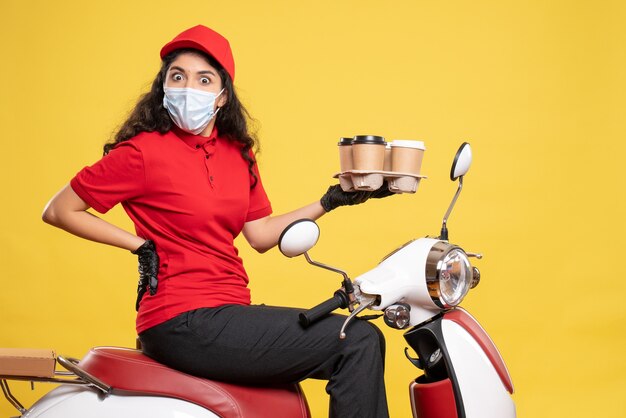Front view female courier in mask on bike with coffee cups on yellow background worker service pandemic uniform job woman covid-