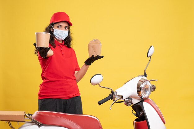 Front view female courier in mask holding delivery dessert and food on a yellow background covid- job uniform worker service delivery