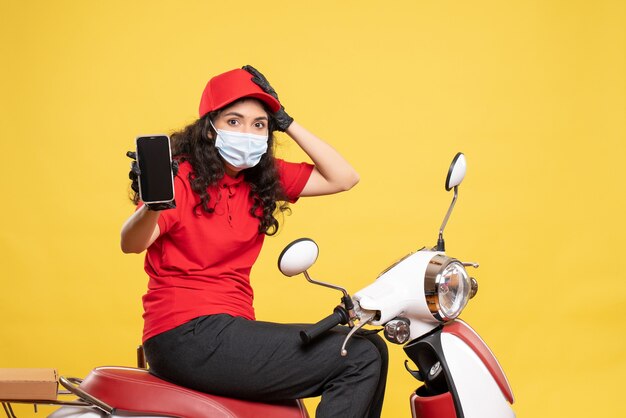 Front view female courier in mask holding phone on the yellow background covid- job uniform worker service pandemic delivery