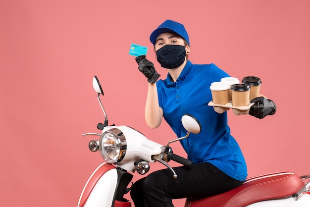 Front view female courier in mask with coffee and bank card on pink 