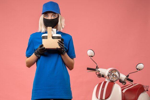 Front view female courier in mask with coffee cups on pink 