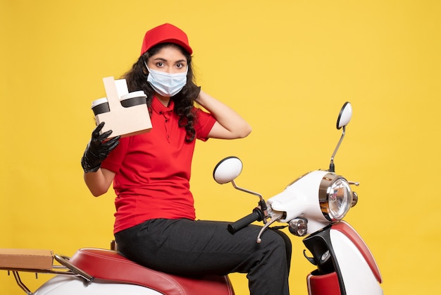 Corriere femminile vista frontale in maschera con tazze di caffè su sfondo giallo servizio covid- uniforme di consegna del lavoro