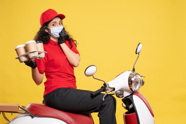 Front view female courier in mask with coffee cups on yellow background service pandemic worker delivery covid- job